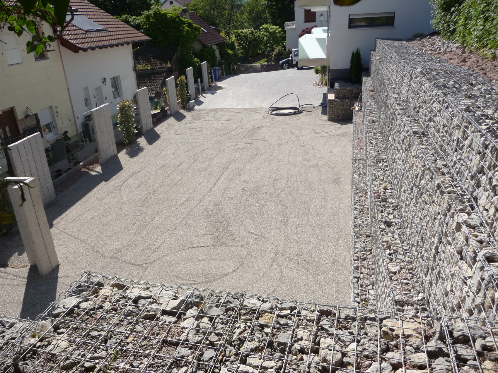 Hofzufahrt mit Kiesvorplatz, Gabionenwand rechts, Betonpfosten links, Häuser im Hintergrund.