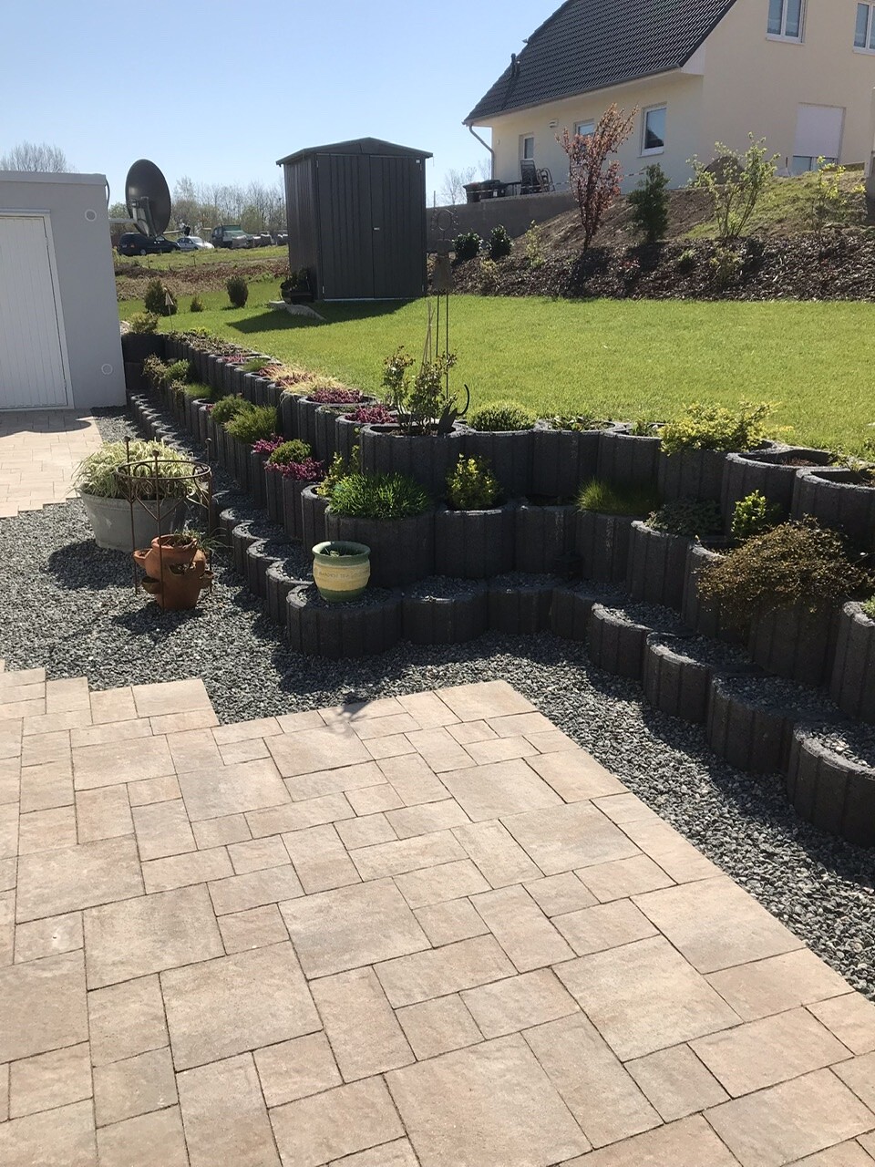 Mehrstufige Beetanlage aus grauen Betonblöcken mit Pflanzen, Terrasse im Vordergrund, Haus im Hintergrund.