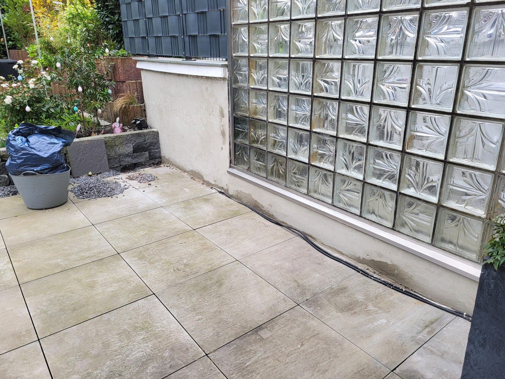 Terrasse mit beigen Fliesen, rechts Glasbausteinwand, links Pflanzkübel mit blauem Beutel.