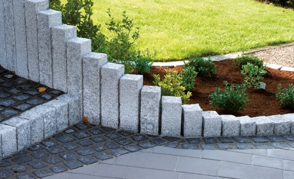 Stufenmauer aus grauen Betonblöcken am Rand eines Pflasterwegs, Garten mit Mulch und Beete.