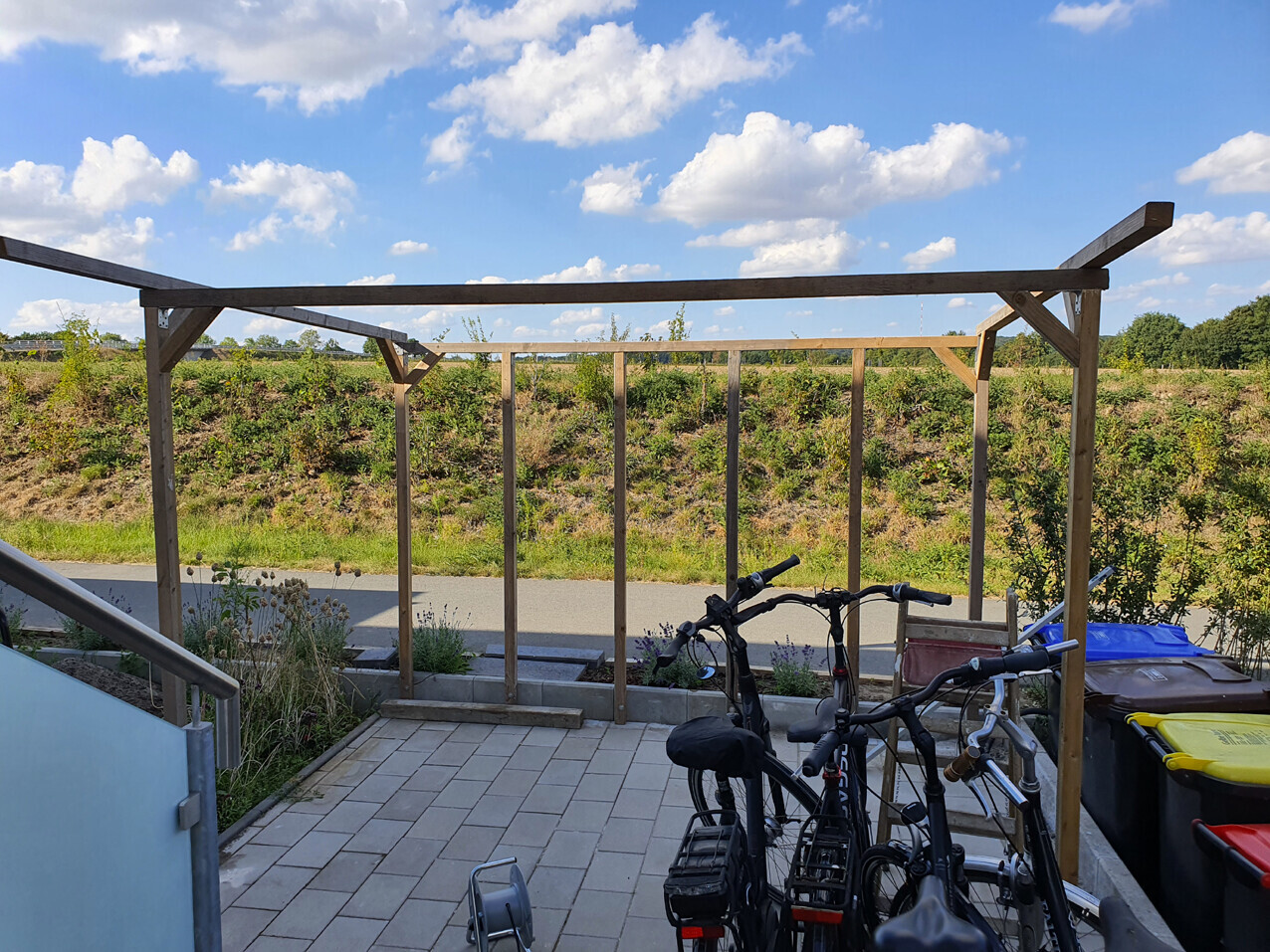 Holzgerüst über einer gepflasterten Terrasse; mehrere Fahrräder davor, Mülltonnen seitlich.