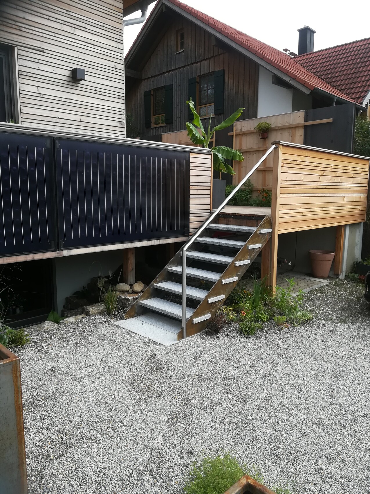 Außentreppe aus Metall führt zu einer Holzterrasse mit Geländer neben dem Haus; Kiesboden und Banane.