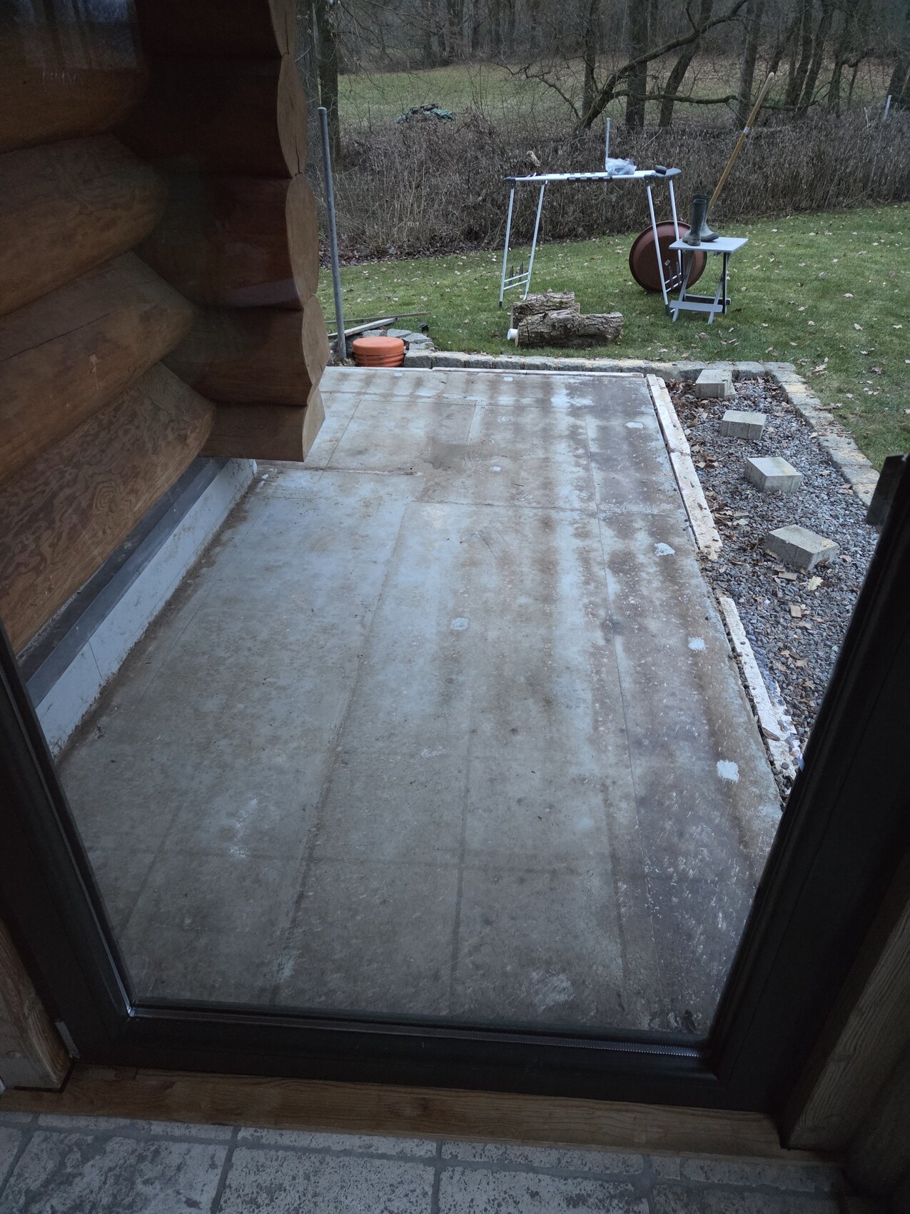Terrasse aus Beton vor einer Holzhütte, Gartenwerkzeug und Baumstämme im Garten