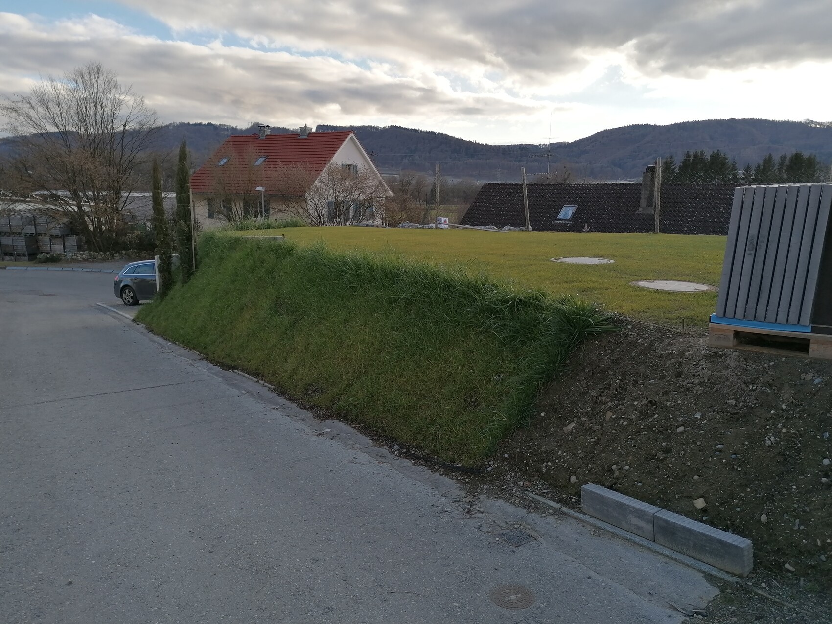 Straßenszene: Grasböschung rechts, Auto links, Haus mit rotem Dach, Berge dahinter.