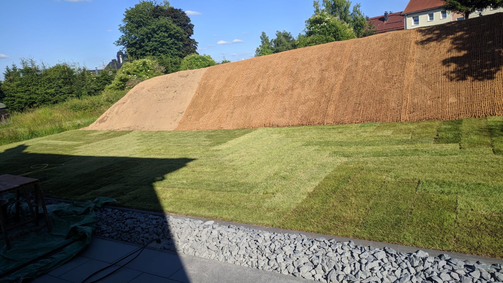 Grüner Garten mit Hang aus Erde und Geotextil, Terrasse und Kiesweg im Vordergrund.