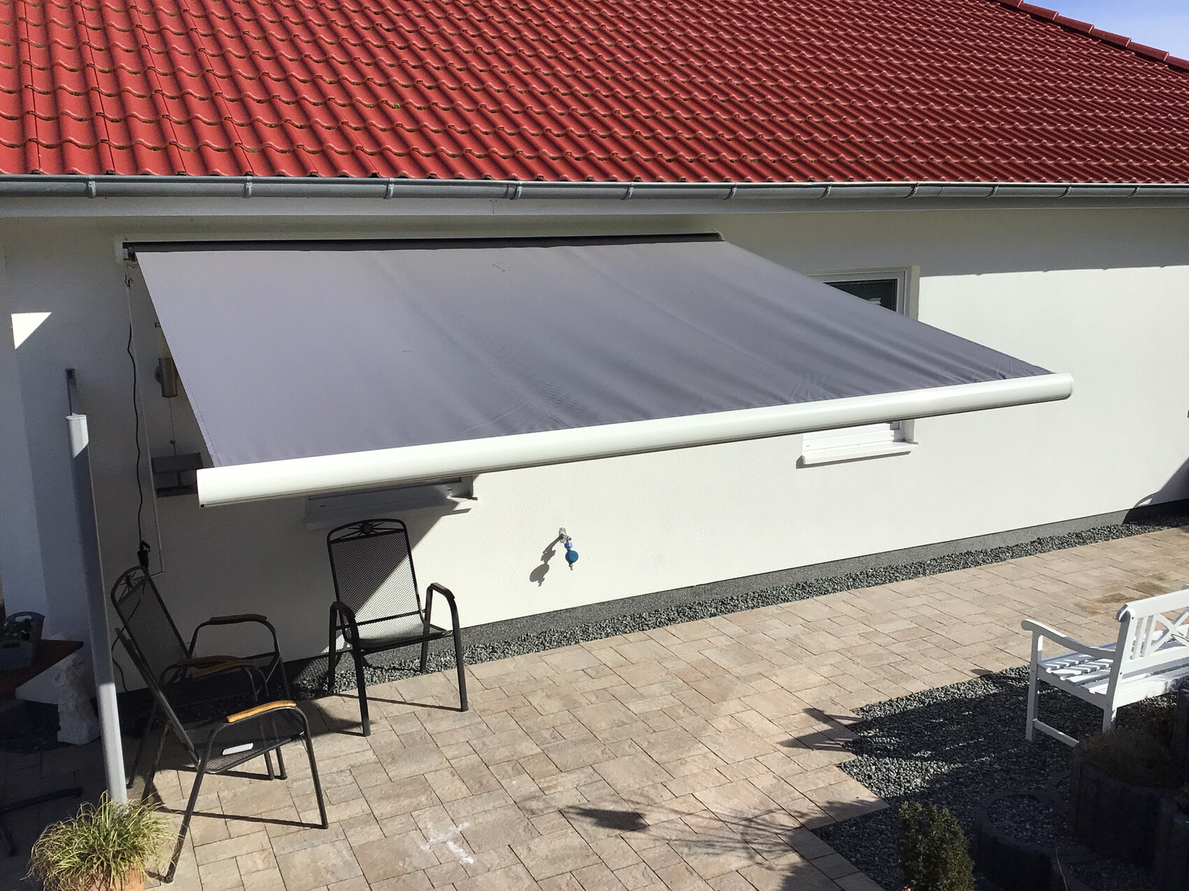 Gepflegter Patio mit grauer Markise über weißer Hauswand, rotem Ziegeldach und Gartenmöbel.