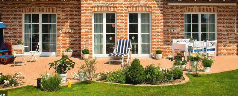 Gartenterrasse vor rotem Backsteinhaus mit Lounge-Sessel, Esstischgruppe und Topfpflanzen.