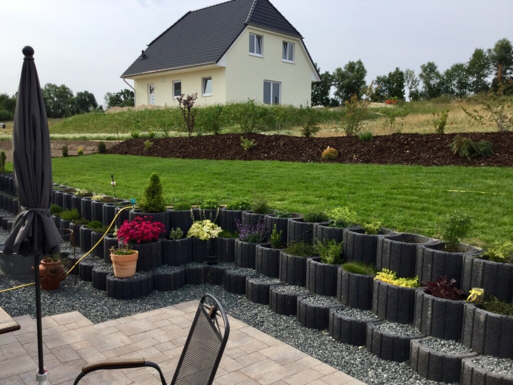 Gartenanlage mit terrassenfoermigen Beetkaesten, gepflasterter Bereich und Haus im Hintergrund.