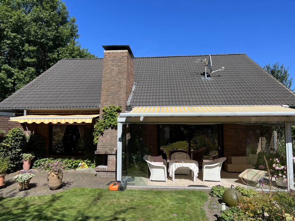 Sonniger Garten mit Terrasse, Hausfront, Kamin, gelb-weiße Markisen und Gartenmobiliar.