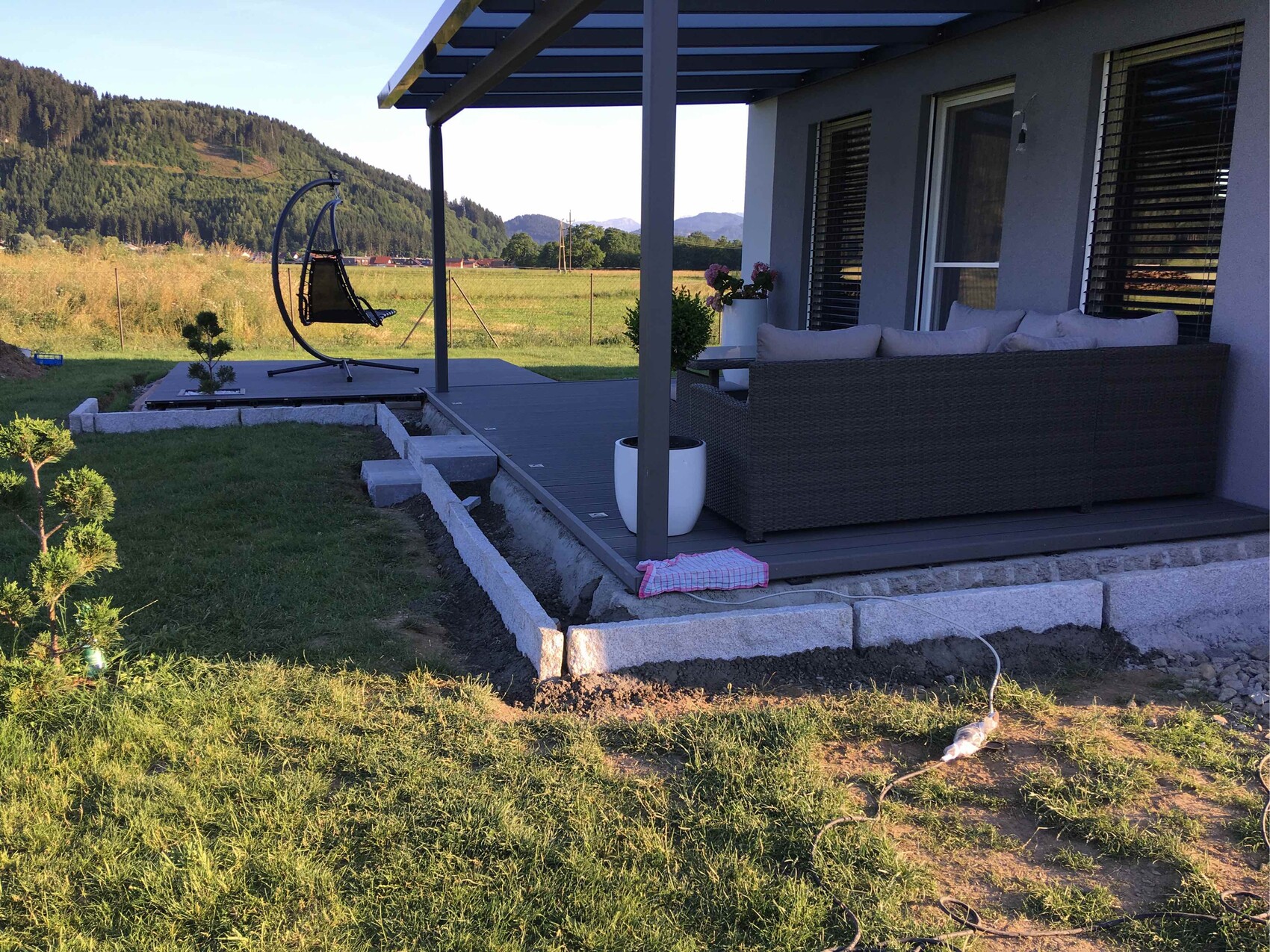Terrasse am Haus mit grauer Outdoor-Sofagruppe und Hängesessel; Berglandschaft im Hintergrund.