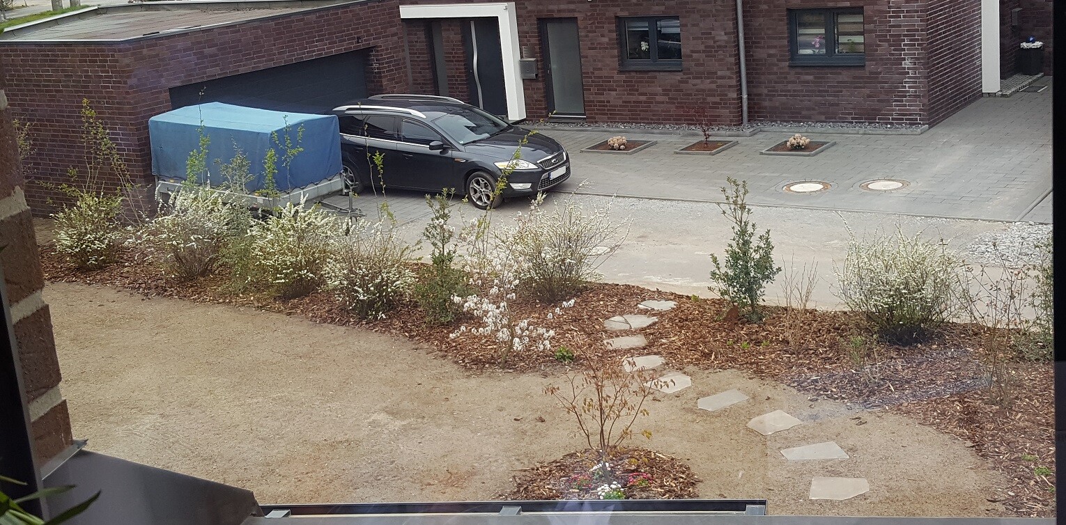 Schwarzer Wagen vor Garage eines Backsteinhauses; blauer Abdeckanhänger, Kiesweg und Sträucher