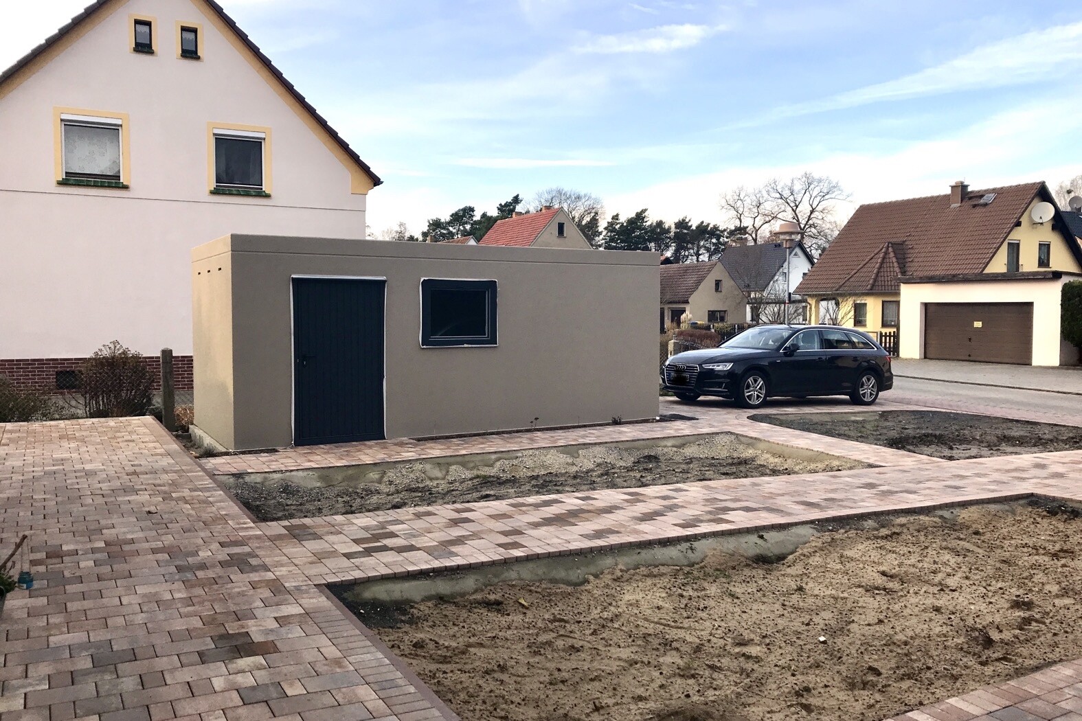 Kleines graues Nebengebäude vor einem beigen Haus; davor ein schwarzes Auto.