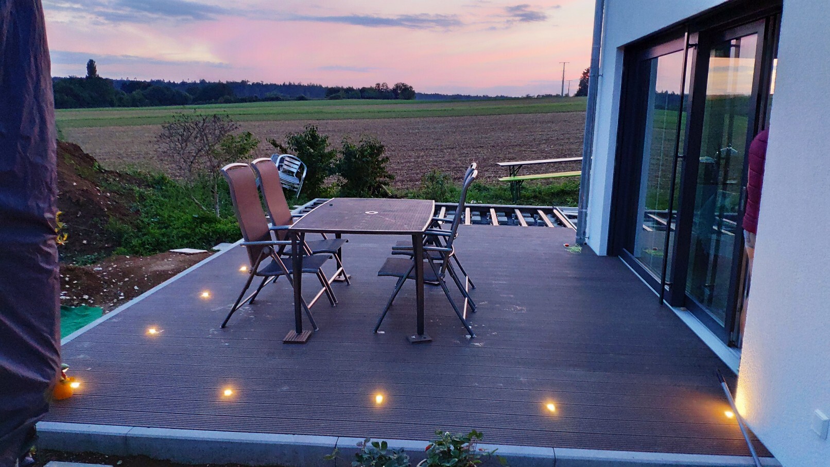 Abendliche Holzterrasse mit Esstisch und Stühlen, Blick aufs Feld hinter dem Haus.