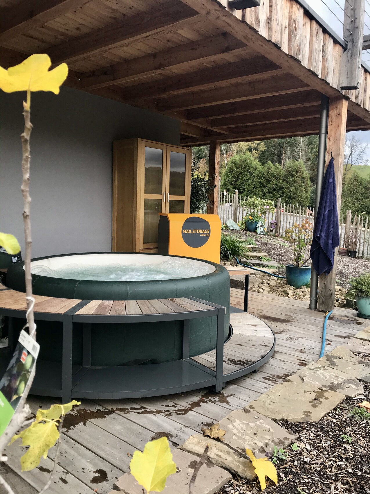 Runder Outdoor-Whirlpool auf Holzdeck unter Pergola; gelbe MAX STORAGE-Box im Hintergrund.