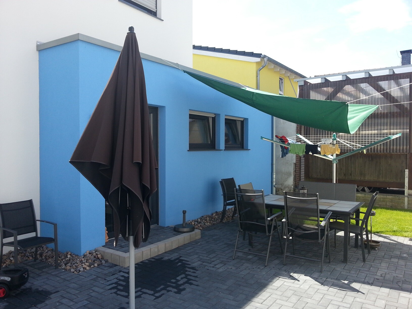 Blick auf blaue Außenwand mit Patio-Tisch und Stühlen, brauner Sonnenschirm und grünem Sonnensegel