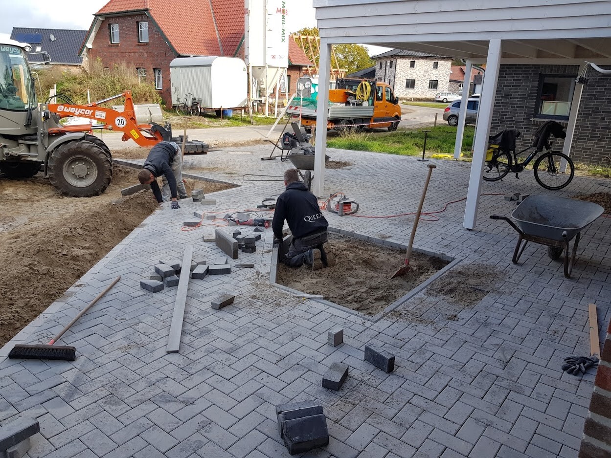 Pflasterarbeiten: zwei Arbeiter legen Pflastersteine, Grube und Bauwerkzeuge auf dem Hof.