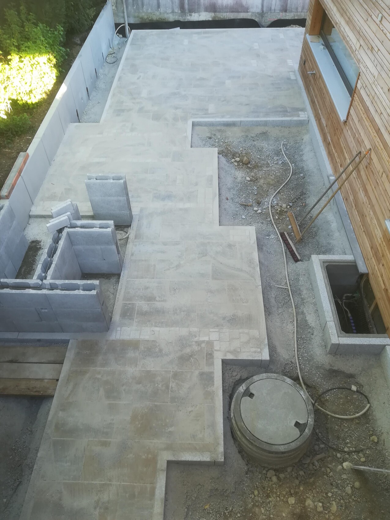 Außenbereich: hellgraue Terrassenplatten in Stufen, Betonblöcke, rechts Holzhauswand, runder Deckel.