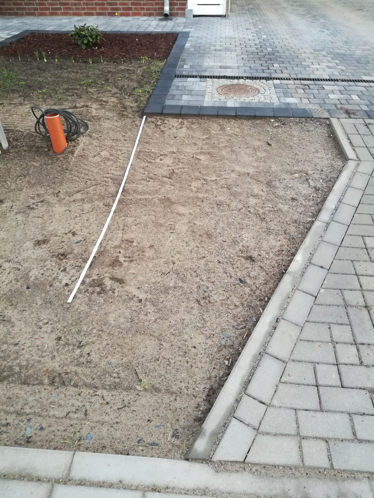 Unbefestigter Erdbereich neben gepflastertem Vorplatz, Kanaldeckel sichtbar