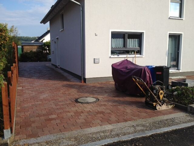 Außenansicht eines Hauses mit gepflasterter Einfahrt; abgedeckte Maschine neben dem Fenster.