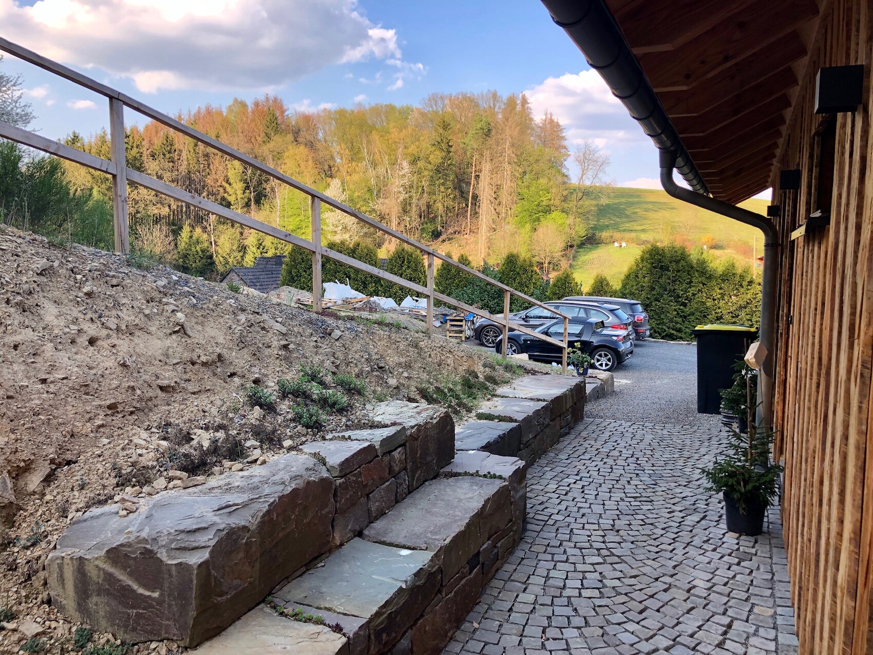 Fußweg neben Holzhaus mit Steinstufen, Geländer, Autos und Wald im Hintergrund.