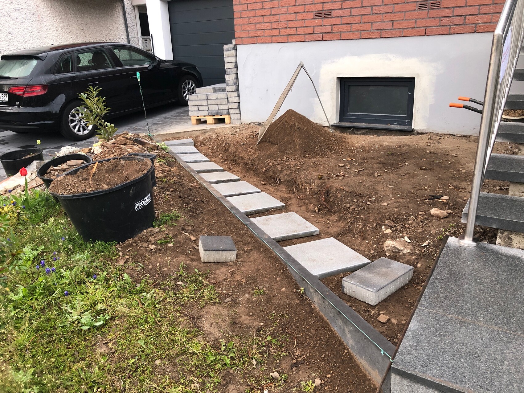 Baustelle vor Haus: Betonplatten als Weg über Erdhügel, Auto im Hintergrund.