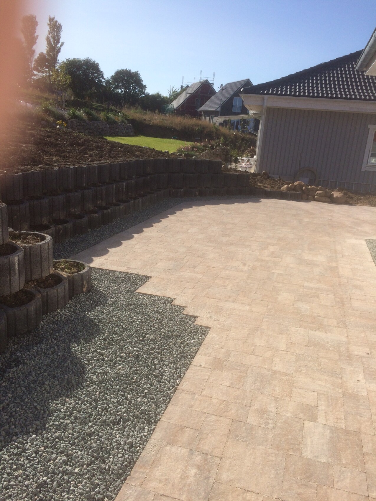 Gepflasterte Terrasse mit Kiesweg, Stützmauer aus Betonsteinen, Haus im Hintergrund.