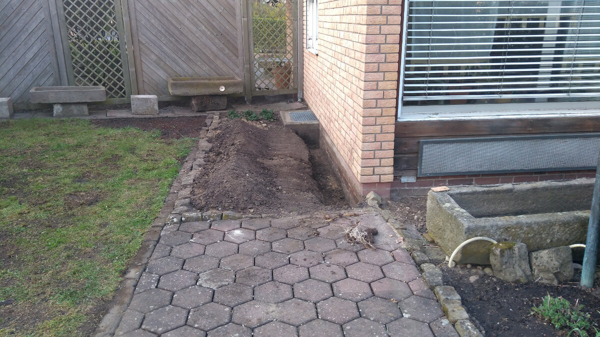 Sechseckige Pflastersteine vor dem Haus; Erdarbeiten entlang der Wand, Holzzaun im Hintergrund.