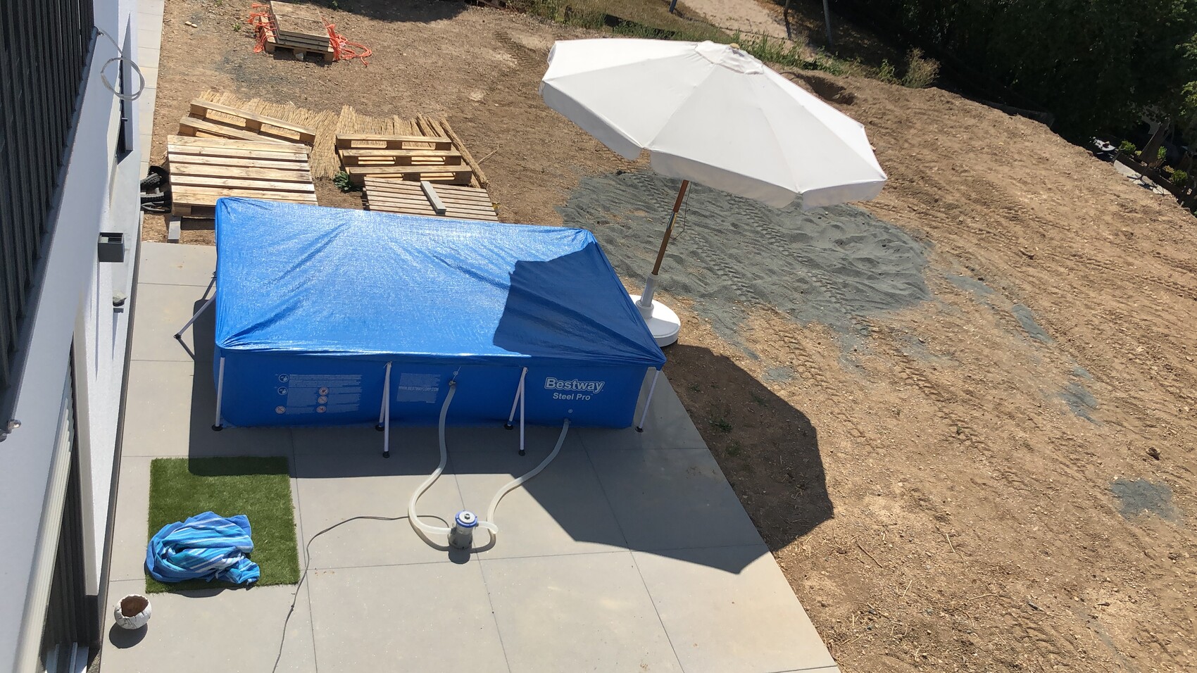 Gartenbau-Baustelle mit blauem Bestway Steel Pro Pool, weißem Sonnenschirm und Paletten.