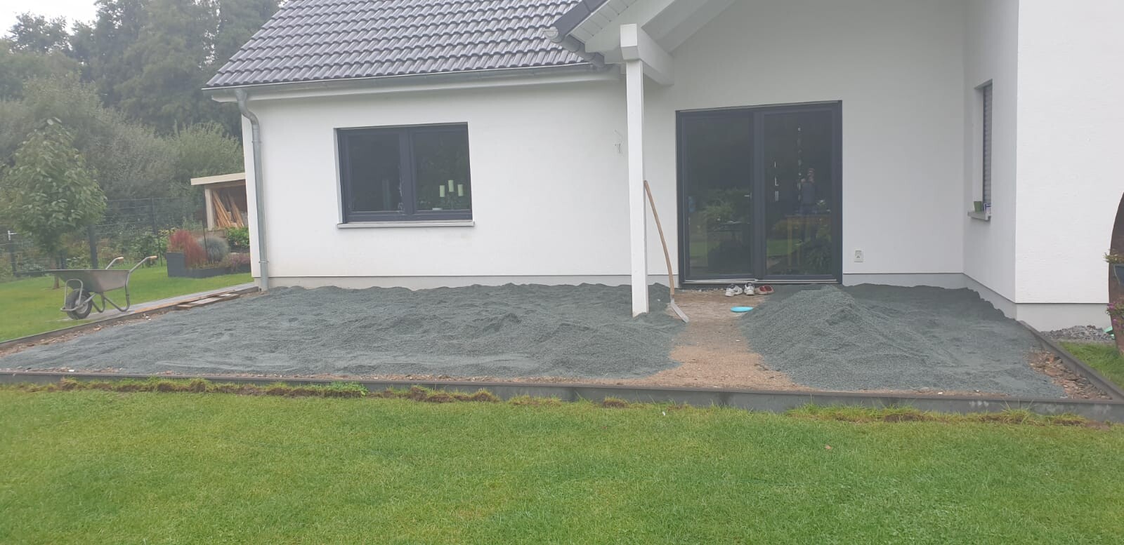 Weiße Hausfassade, Kies-Terrassenboden wird vorbereitet; Glastür und Fenster, linke Schubkarre.