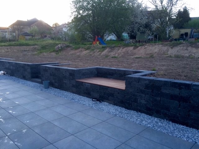Dunkle Steinmauer mit eingebauter Holzbank vor einer gepflasterten Terrasse, Garten im Hintergrund.