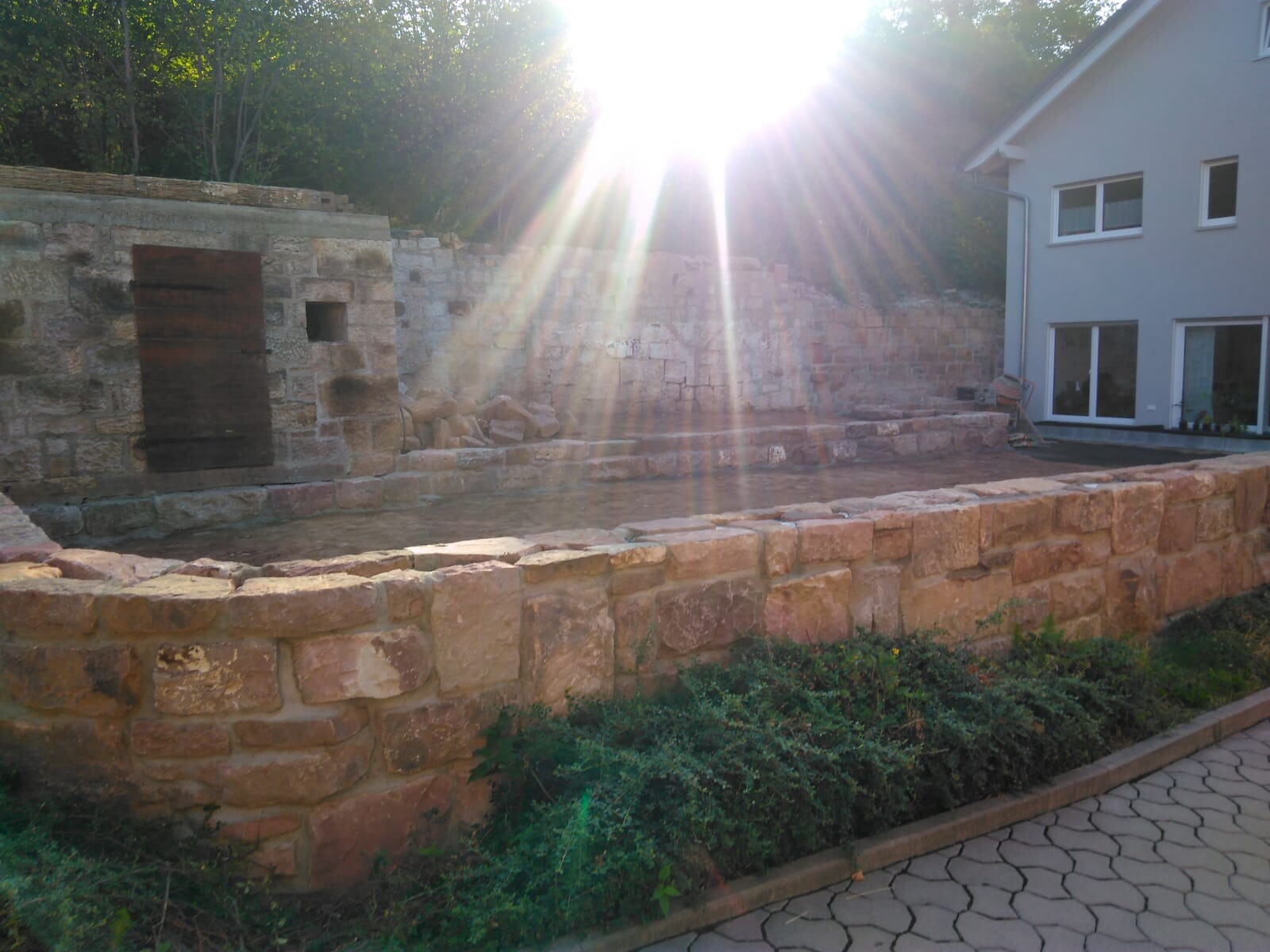 Außenbereich mit Steinmauer, Terrasse und modernem Haus im Hintergrund bei Sonnenschein