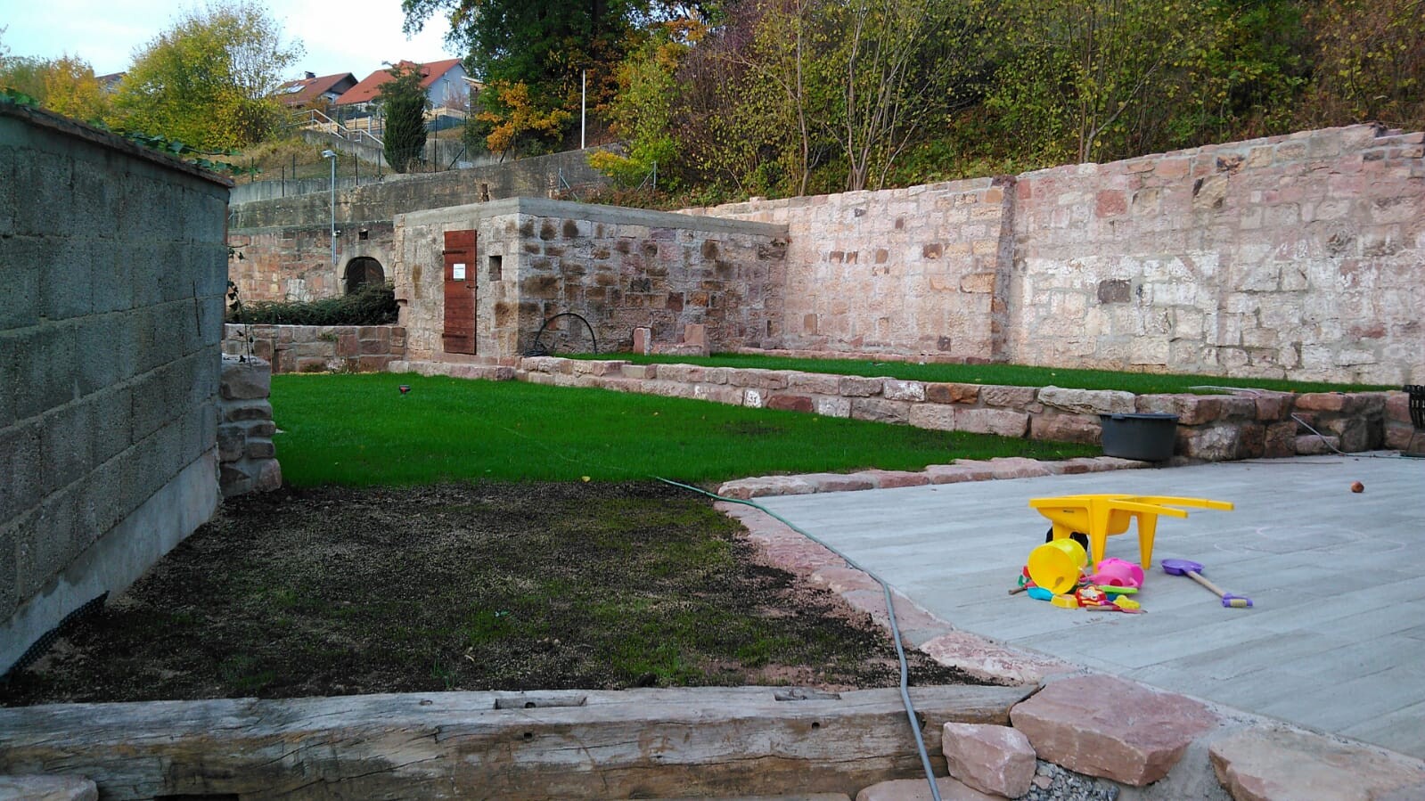 Hinterhof mit Steinmauer, grünem Rasen und buntem Spielzeug auf Betonfläche