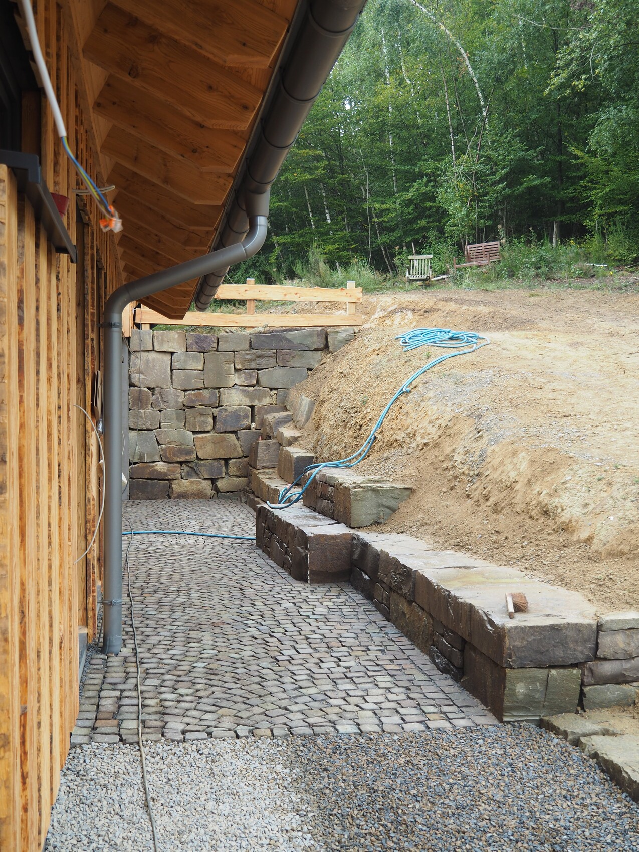 Außenansicht einer Baufläche mit Steinpflaster-Terrasse, Fallrohr und Wald im Hintergrund