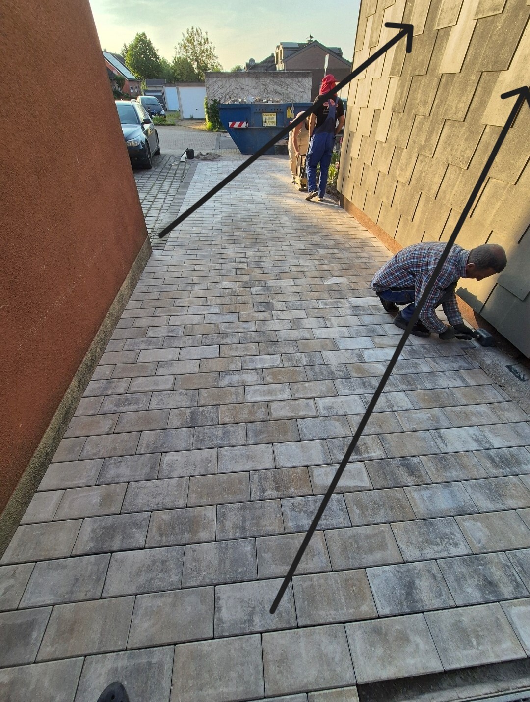 Arbeiter verlegen Pflastersteine neben einer Hauswand; Baustellencontainer im Hintergrund.