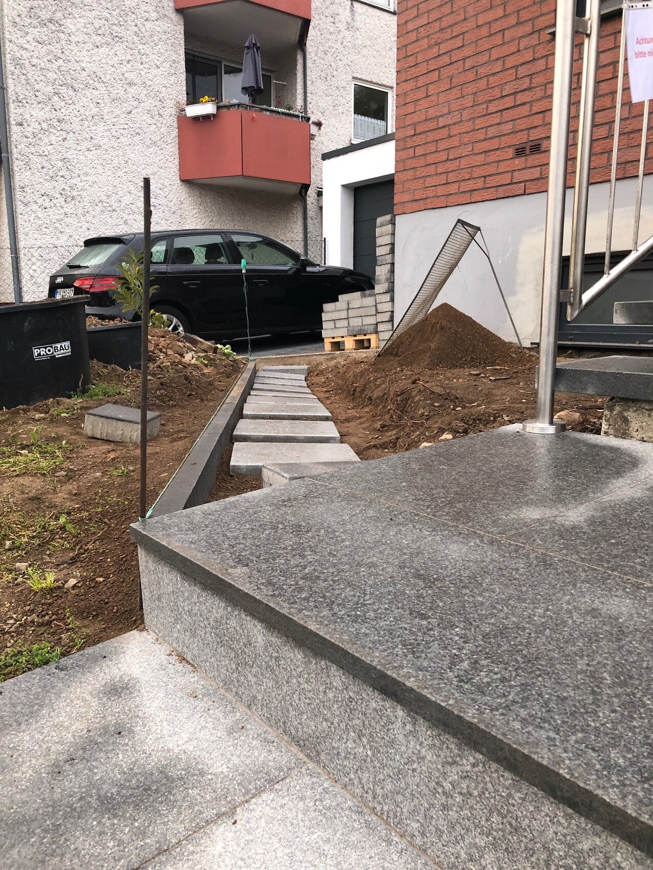 Baustelle vor Wohnhaus: Stein-Stufenweg führt zum Eingang; Kieshaufen, Paletten, schwarzes Auto.