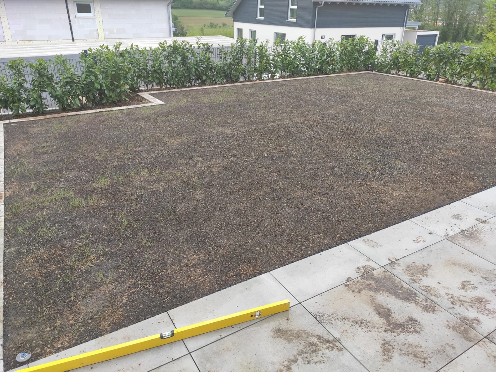 Große braune Erdfläche auf Terrasse, Rand mit Hecke; gelbe Wasserwaage liegt auf Pflaster.