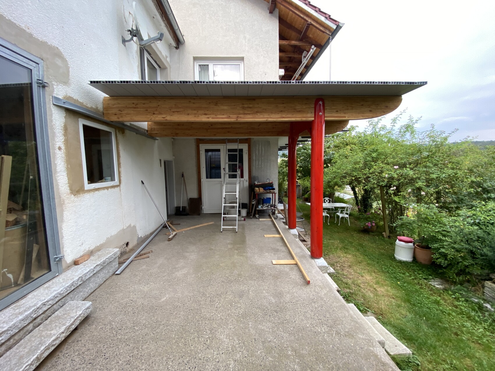 Überdachte Terrasse mit Holzbalken, roter Säule, Leiter und Bauwerkzeugen; Garten dahinter.