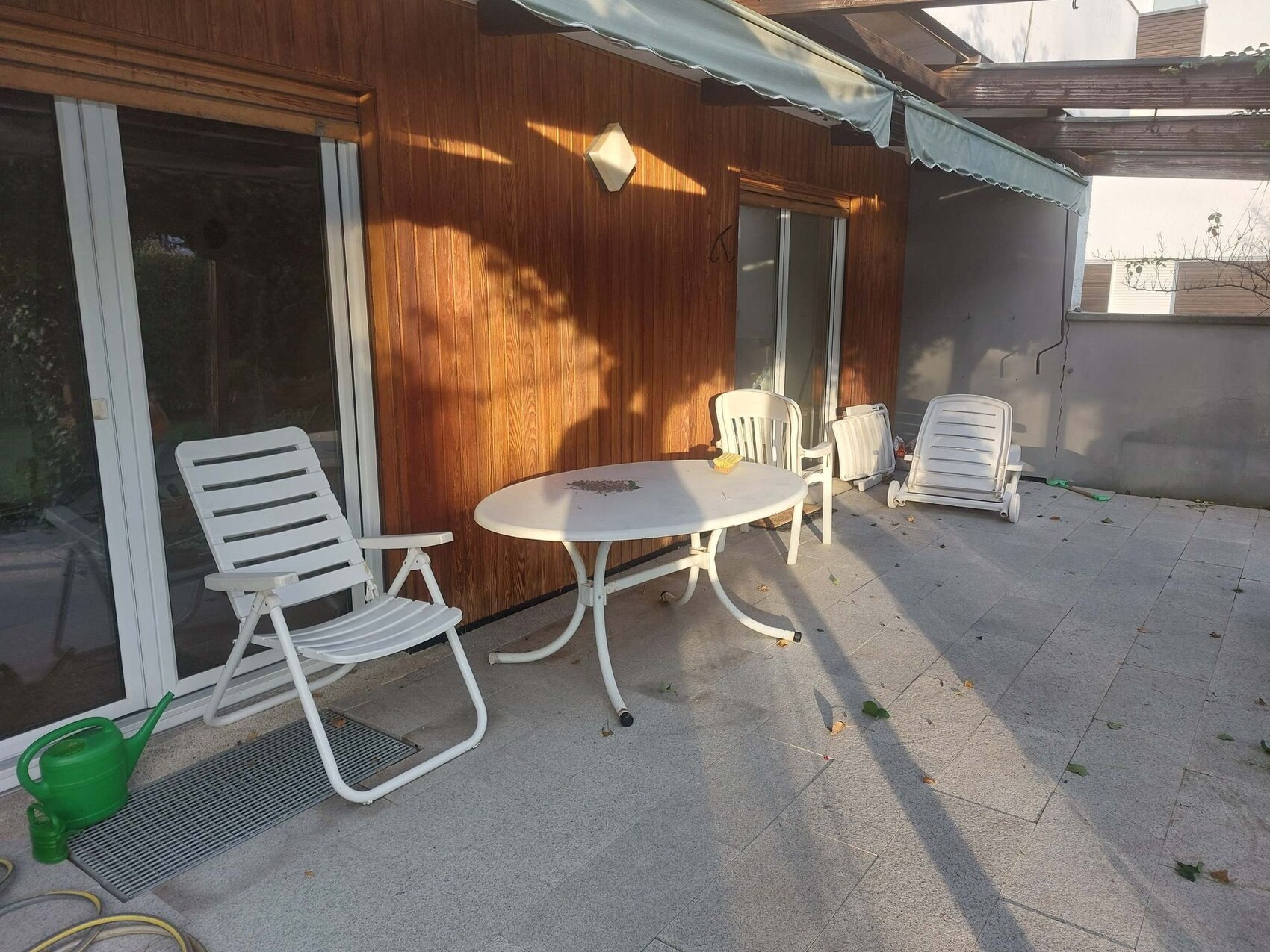 Terrasse mit weißen Kunststoffstühlen, runder Tisch und Liegen vor Holzwand.