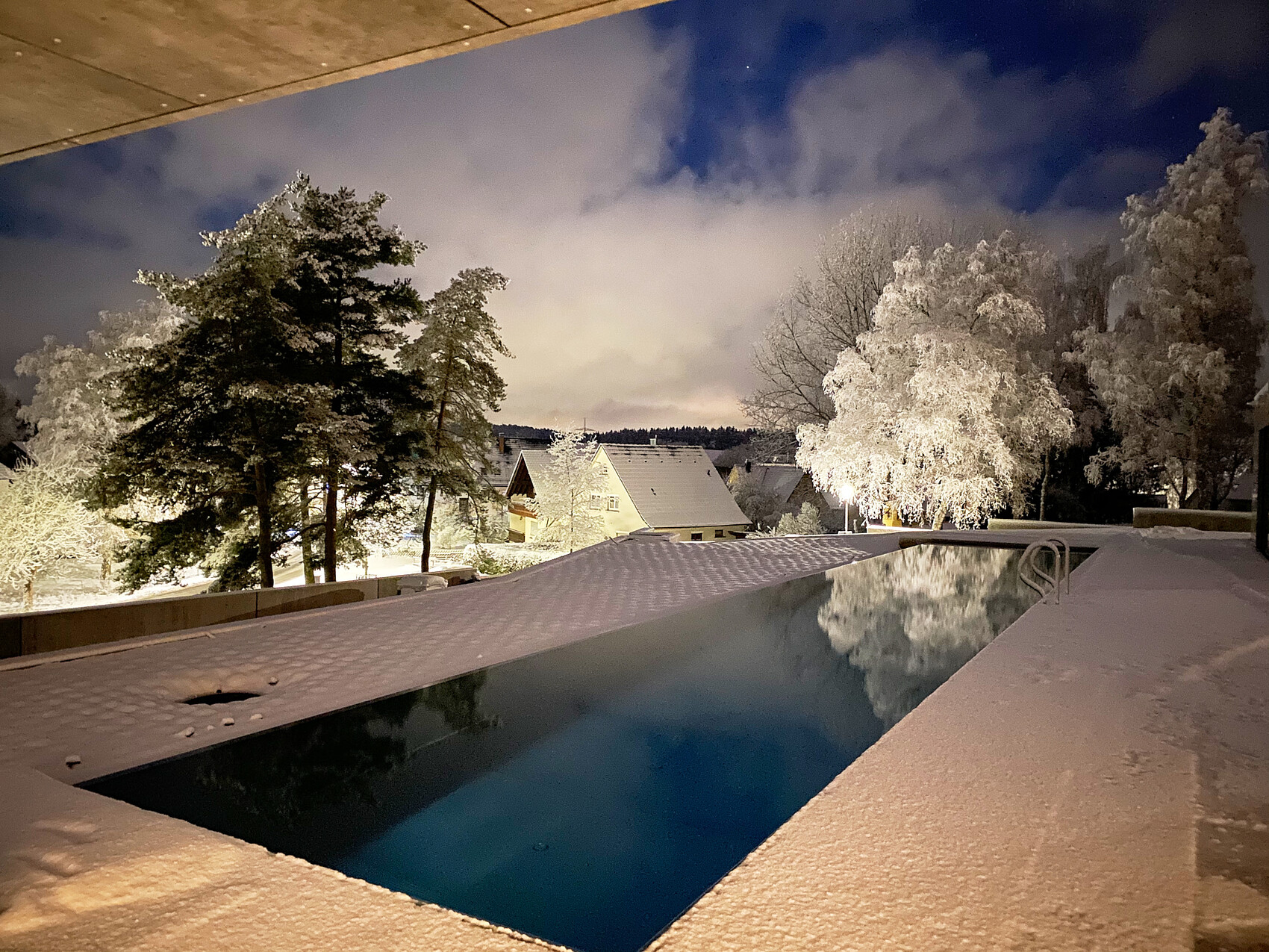 Nachtaufnahme einer verschneiten Terrasse mit Pool, Bäumen und Häusern im Hintergrund.