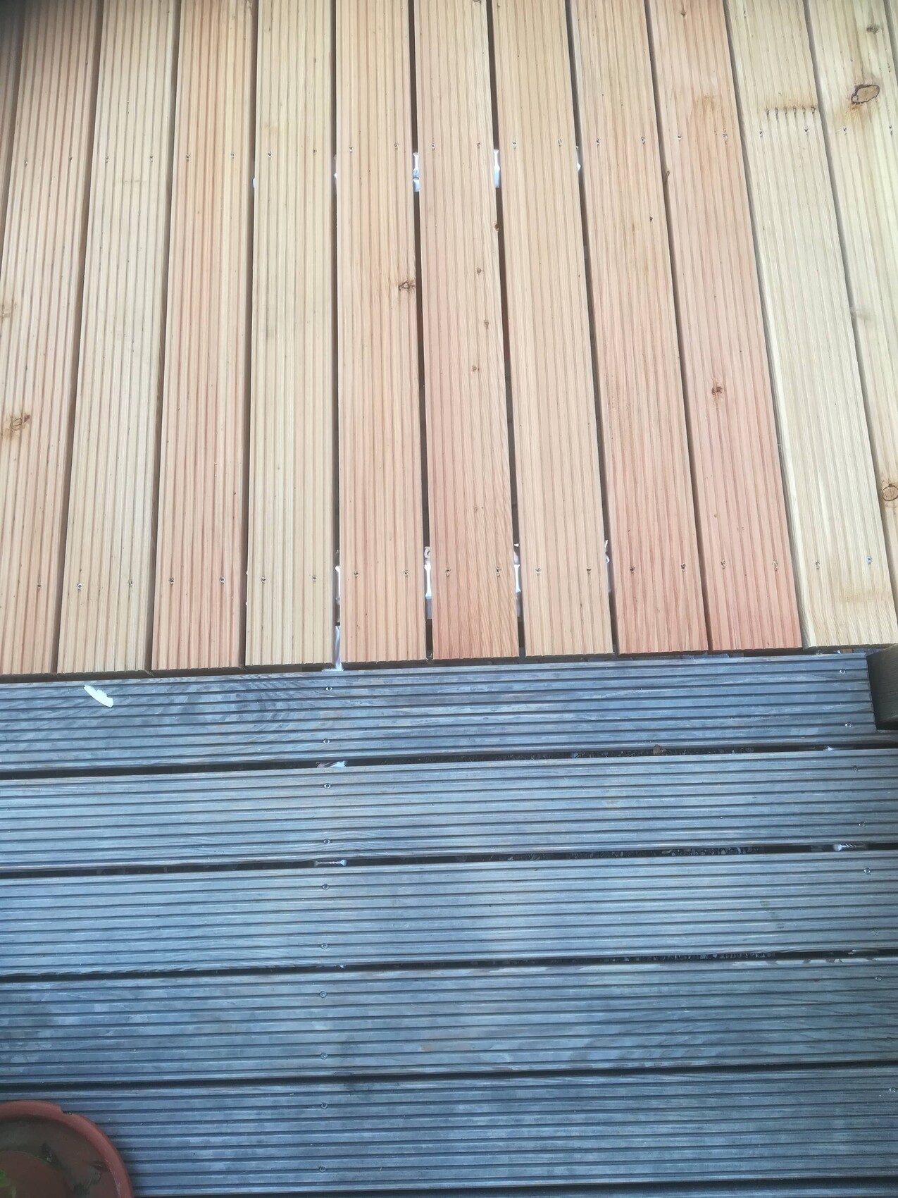 Holzdeck mit vertikalen Dielen oben; darunter grau gerillte Planken, linker Topf sichtbar.
