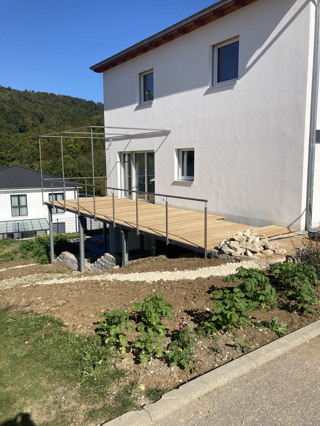 Weißes zweistöckiges Haus mit Holzterrasse, Metallgeländer und Gartenpflanzen im Vordergrund.