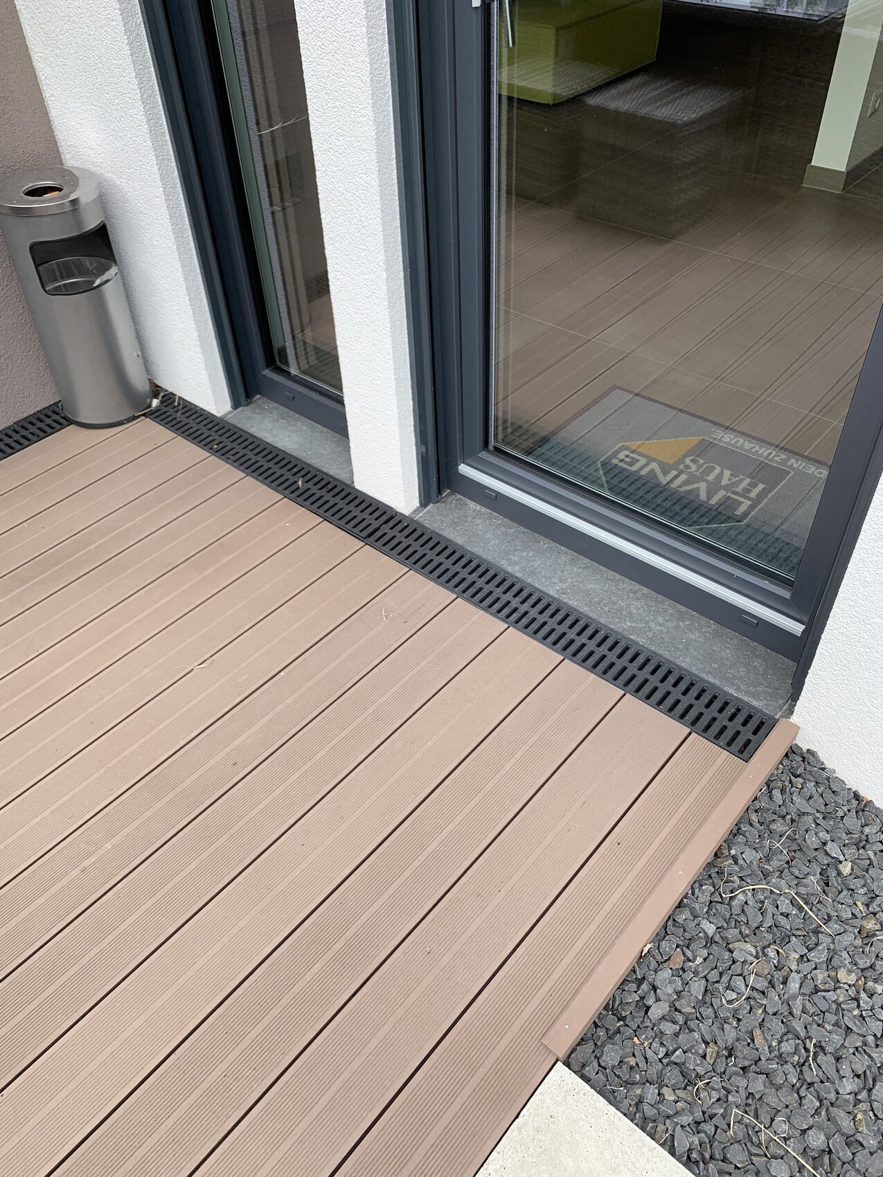 Balkon mit braunem Holzdeck, Glasschiebetür, schwarzer Drainageleiste und Mülleimer links.