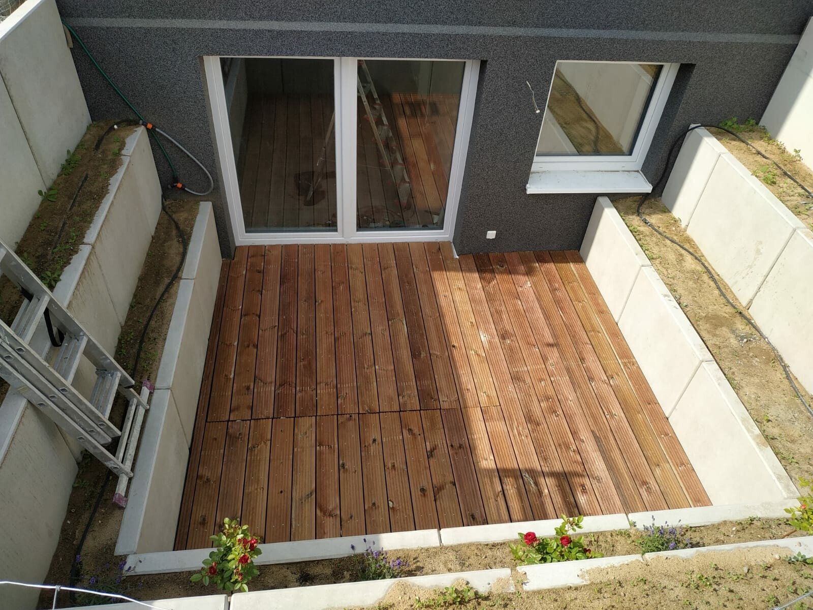 Blick von oben auf eine Holzdeck-Terrasse vor einem Haus, mit Glasschiebetür und Betonwänden.