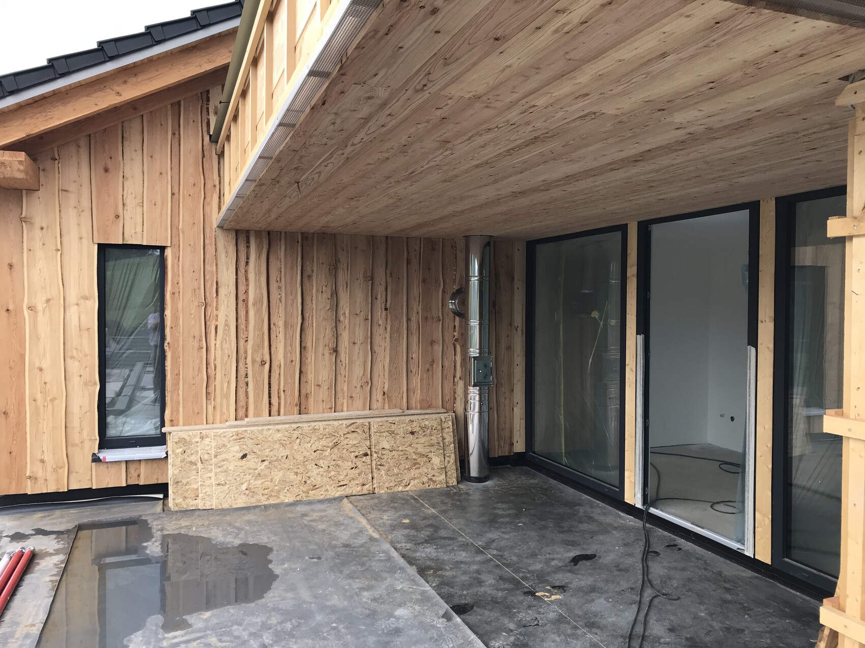 Außenansicht eines Holzhauses im Bau mit vertikaler Holzverkleidung, Glas-Schiebetüren und Metallrohr.