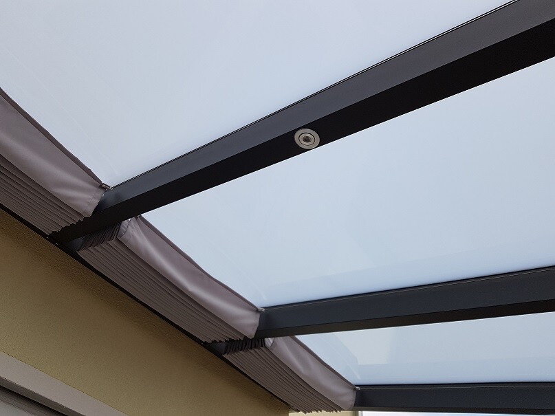 Überdachte Terrasse mit Glasdach, schwarzen Trägern und Stoffmarkisen; kleine Kamera am Träger.