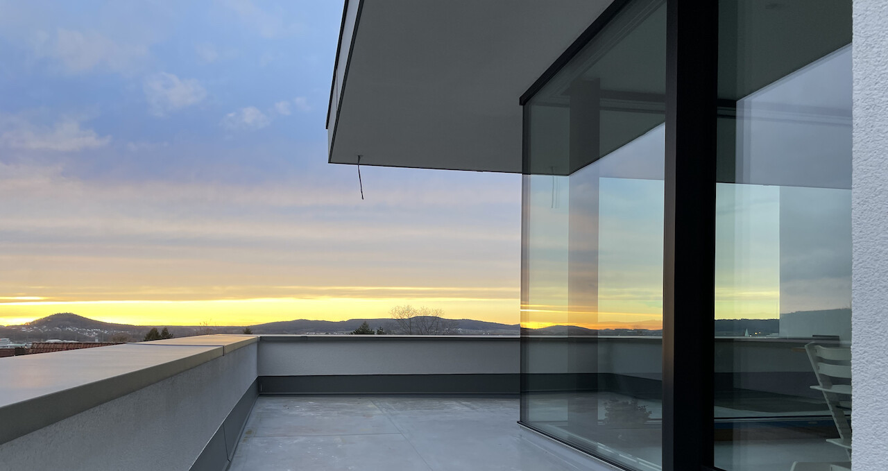 Moderne Dachterrasse mit Glasfront, Blick auf Sonnenuntergang über Hügeln.