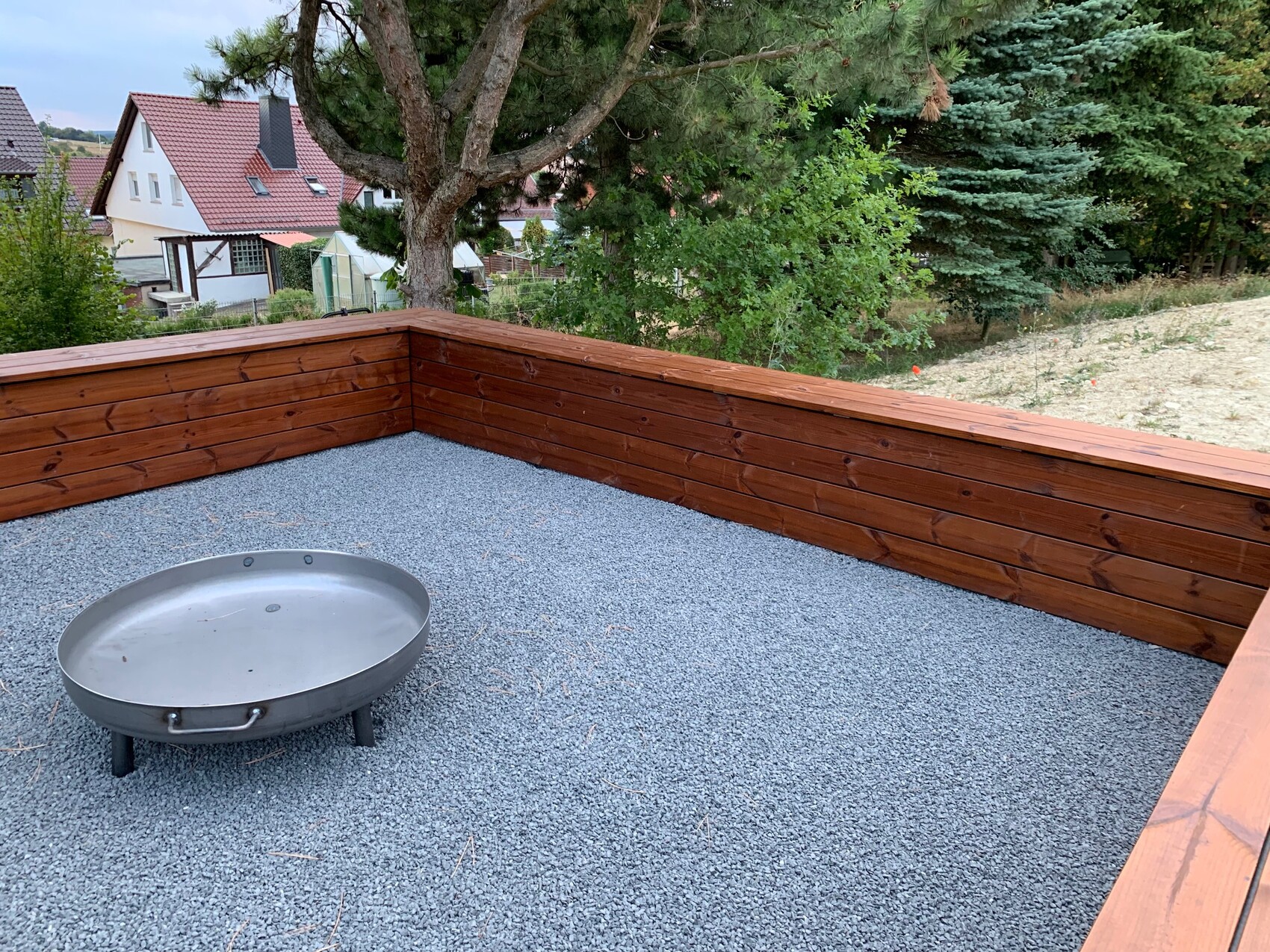 Terrasse mit Holzverkleidung, Kiesboden, runder Feuerkorb, Bäume und Nachbarhäuser im Hintergrund.