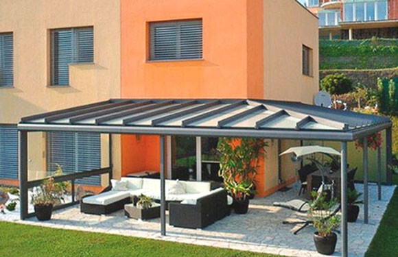 Außenbereich unter moderner Glaspergola mit Glasdach, Lounge-Möbel, Pflanzen und Grill.