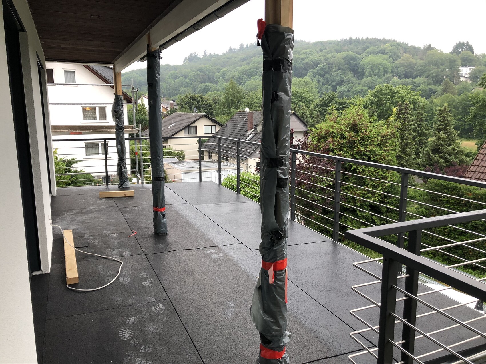 Balkon mit drei Stützen, in Folie eingewickelt; Blick auf grüne Vororte und Wald.