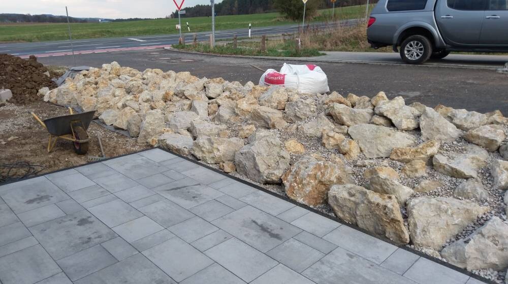 Großer Steinwall neben frisch gepflasterter Terrasse; Schubkarren, Sack und Pkw im Hintergrund.